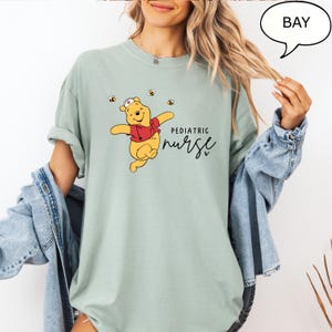 May include: A light green t-shirt featuring a cartoon Winnie the Pooh character wearing a nurse's hat and red shirt, with the words "Pediatric Nurse" in black script. Three small yellow bees are also depicted on the shirt.