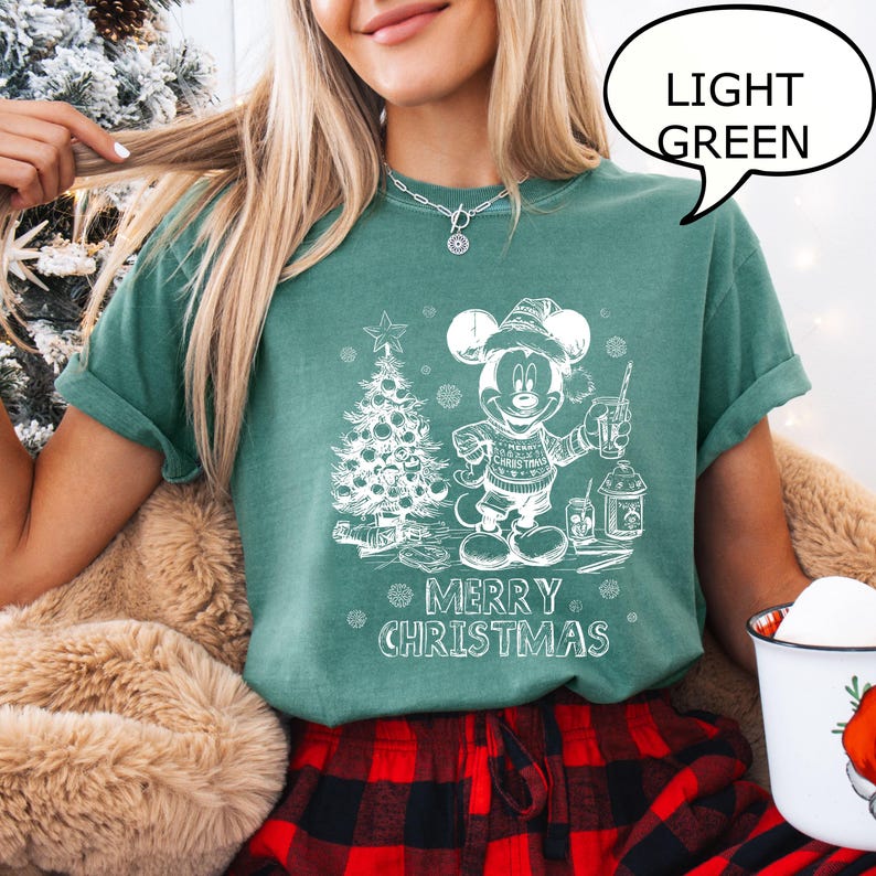 Puede incluir: Camiseta verde claro con un gr&aacute;fico blanco de Mickey Mouse con un su&eacute;ter navide&ntilde;o, un &aacute;rbol de Navidad y las palabras "Merry Christmas". La camiseta se combina con pantalones de pijama a cuadros rojos y negros.