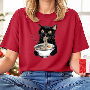 Puede incluir: Una camiseta roja con un gato negro comiendo fideos ramen. El gato sostiene un taz&oacute;n blanco con palillos que sobresalen. El gato tiene una expresi&oacute;n de sorpresa en su rostro.