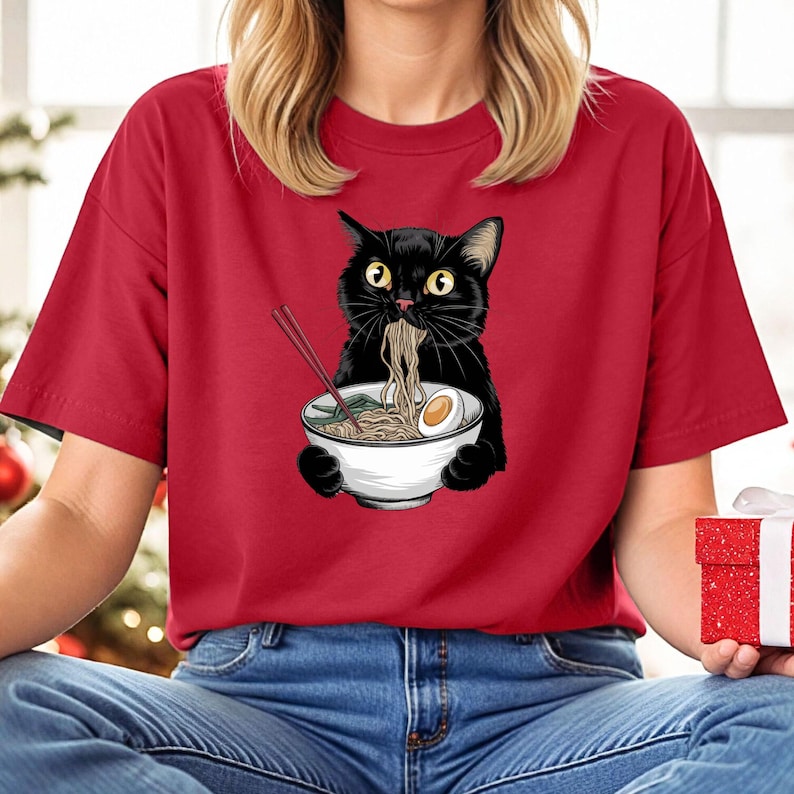 Puede incluir: Una camiseta roja con un gato negro comiendo fideos ramen. El gato sostiene un taz&oacute;n blanco con palillos que sobresalen. El gato tiene una expresi&oacute;n de sorpresa en su rostro.