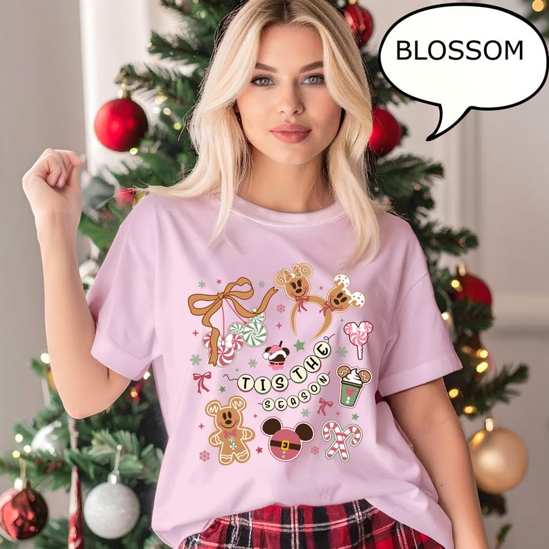May include: A light pink t-shirt with a festive design featuring gingerbread men, candy canes, and the text "'Tis the Season". The design includes Mickey Mouse-shaped elements and a bow with candy. The word "BLOSSOM" is in a speech bubble.