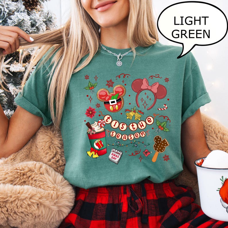 May include: Light green t-shirt with a festive graphic featuring Mickey Mouse-themed Christmas elements. The design includes a gingerbread cookie, a bow, a coffee cup, and the text "'Tis the Season". The shirt is paired with red and black plaid pajama bottoms.