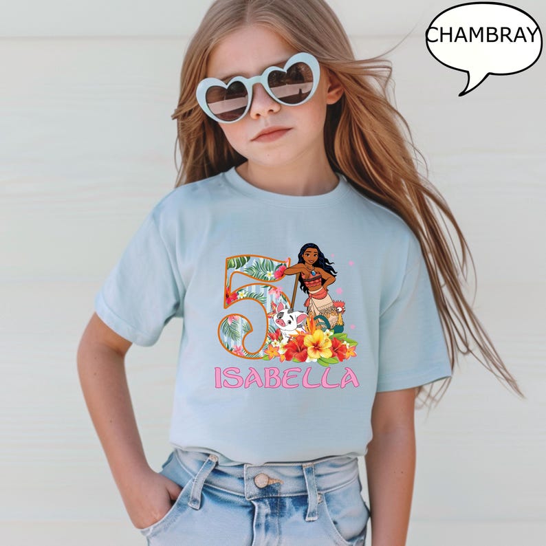 May include: A light blue t-shirt featuring a colorful graphic of a cartoon character, flowers, and the number 5. The name "ISABELLA" is printed in pink below the graphic. The word "CHAMBRAY" is in a speech bubble.