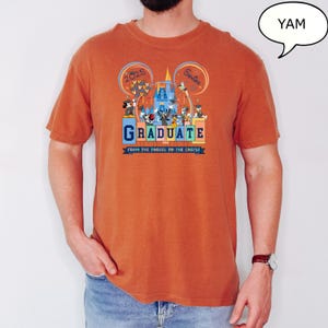 以下が含まれることがあります： さび色のTシャツに、ミッキーマウスとその仲間たちがディズニーワールドで卒業を祝っているグラフィックがプリントされています。Tシャツの文字は「2025 Senior Graduate From the Tassel to the Castle」です。