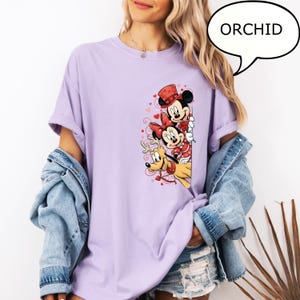 Puede incluir: Una camiseta de color lila claro con un gr&aacute;fico de Mickey Mouse, Minnie Mouse y Pluto. Mickey y Minnie visten trajes rojos y blancos. La palabra "ORCHID" est&aacute; en un bocadillo.
