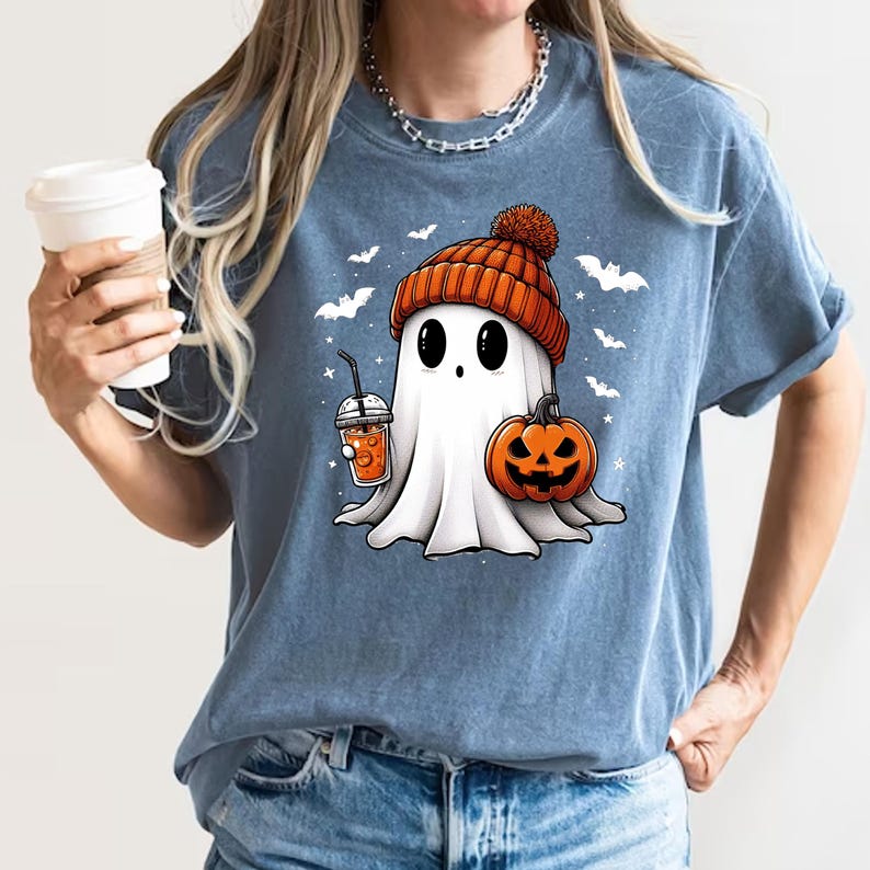 May include: A blue t-shirt featuring a cartoon ghost wearing a pumpkin-coloured beanie and holding a pumpkin and a cup of orange liquid. The ghost has a cute face with big eyes and a small mouth. There are bats flying around the ghost.