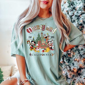 Puede incluir: Una camiseta verde claro con un gr&aacute;fico de Disneyland con tem&aacute;tica navide&ntilde;a. El gr&aacute;fico presenta a Mickey Mouse, Donald Duck, Goofy y Pluto frente al Castillo de la Bella Durmiente. El texto "Disneyland Est. 1955 California" est&aacute; impreso en la camiseta.