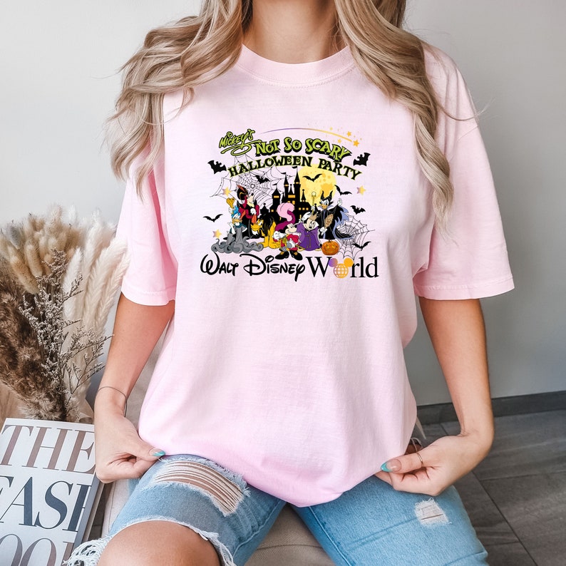 May include: A pink t-shirt with a graphic print of Mickey Mouse and other Disney characters dressed in Halloween costumes. The text on the shirt reads "Mickey's Not So Scary Halloween Party Walt Disney World".