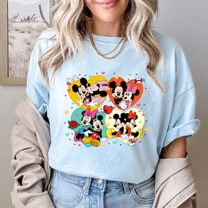 Puede incluir: Una camiseta azul claro con un estampado gr&aacute;fico de Mickey Mouse y Minnie Mouse en cuatro dise&ntilde;os diferentes en forma de coraz&oacute;n. Los corazones est&aacute;n llenos de lunares rojos, amarillos y rosas.