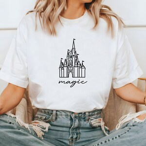May include: White t-shirt with a black outline of a castle and the word "magic" written underneath.