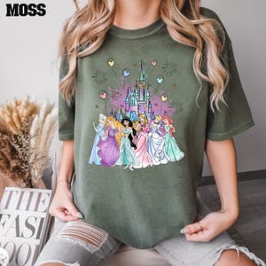 Puede incluir: Una camiseta verde con un estampado gr&aacute;fico de princesas Disney frente al castillo de Cenicienta. Las princesas llevan vestidos coloridos y hay globos y fuegos artificiales en el fondo.