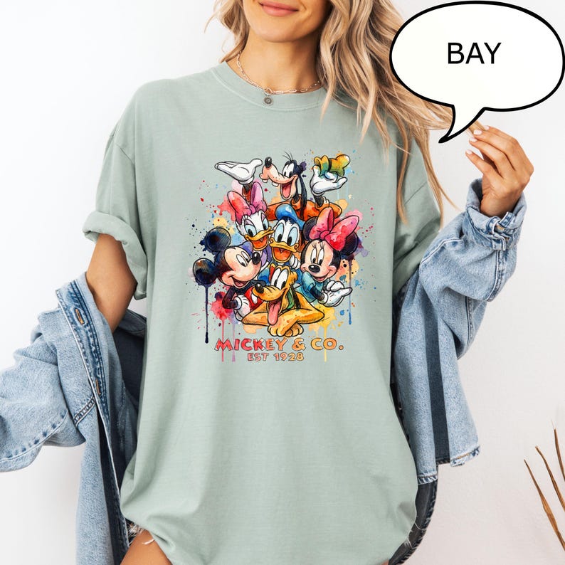 May include: A light green t-shirt featuring a colorful graphic of Mickey Mouse and friends. The design includes Mickey, Minnie, Donald, Daisy, Goofy, and Pluto, with the text "MICKEY & CO. EST 1928". The shirt has a relaxed fit.