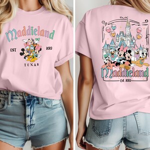 K&ouml;nnte beinhalten: Rosa T-Shirt mit einem Aufdruck von Mickey Mouse und seinen Freunden vor einem Schloss. Der Text "Maddieland Est. 2023" befindet sich auf dem Shirt.