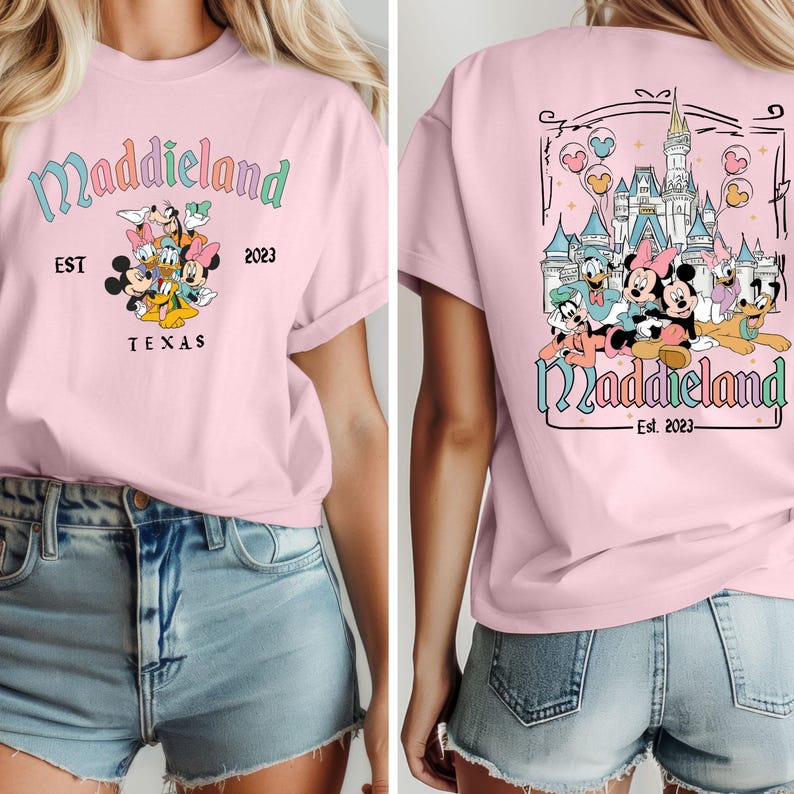 K&ouml;nnte beinhalten: Rosa T-Shirt mit einem Aufdruck von Mickey Mouse und seinen Freunden vor einem Schloss. Der Text "Maddieland Est. 2023" befindet sich auf dem Shirt.
