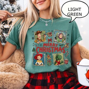 May include: Light green t-shirt featuring a "Merry Christmas" design with cartoon characters. The design includes images of Woody, Buzz Lightyear, Jessie, and Rex. The shirt is paired with red and black plaid pajama pants.
