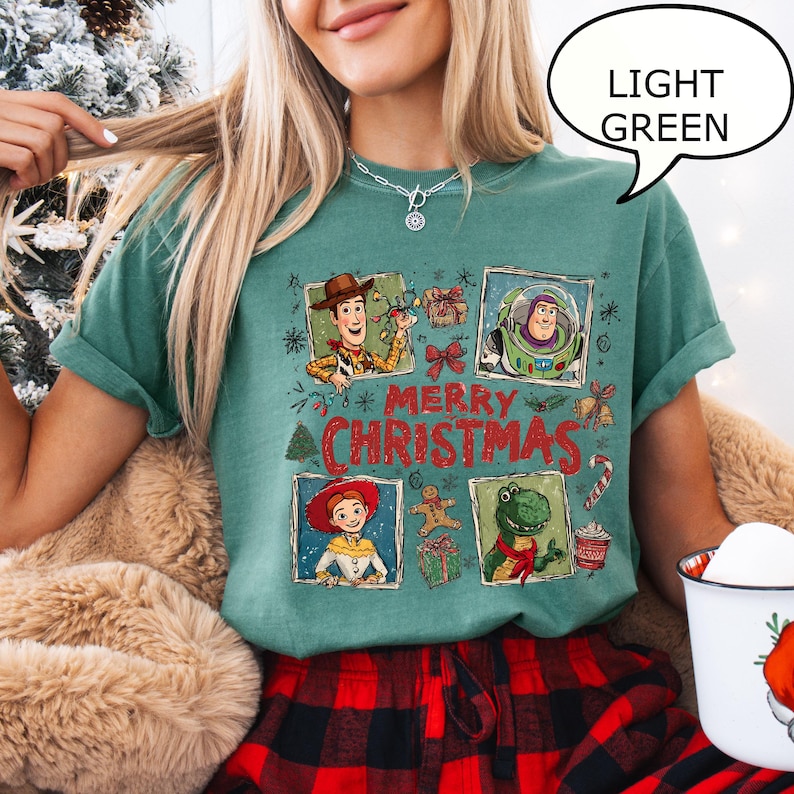 May include: Light green t-shirt featuring a "Merry Christmas" design with cartoon characters. The design includes images of Woody, Buzz Lightyear, Jessie, and Rex. The shirt is paired with red and black plaid pajama pants.