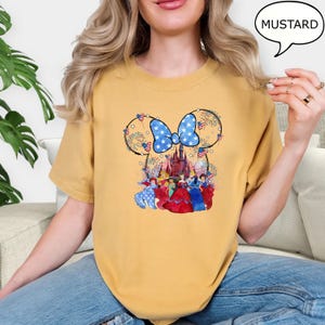 Puede incluir: Camiseta color mostaza con un dise&ntilde;o de Minnie Mouse, un lazo azul y un castillo. El dise&ntilde;o incorpora fuegos artificiales y elementos patri&oacute;ticos. La camiseta tiene un ajuste c&oacute;modo.