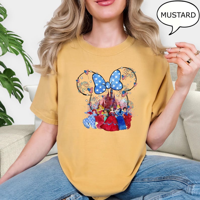 Puede incluir: Camiseta color mostaza con un dise&ntilde;o de Minnie Mouse, un lazo azul y un castillo. El dise&ntilde;o incorpora fuegos artificiales y elementos patri&oacute;ticos. La camiseta tiene un ajuste c&oacute;modo.
