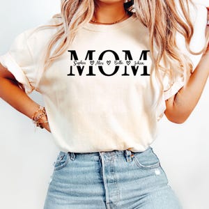 May include: Cream-colored t-shirt with the word "MOM" in bold black letters. Below the word "MOM" are the names Sophia, Alex, Belle, and Johan, each separated by a heart symbol. The shirt is made of a soft material.