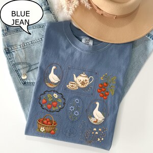 Puede incluir: Una camiseta azul con un estampado gr&aacute;fico de varias ilustraciones que incluyen gansos, una tetera con taza, fresas en una cesta, fresas sueltas, flores azules y tomates rojos, todo ello dispuesto en marcos decorativos.