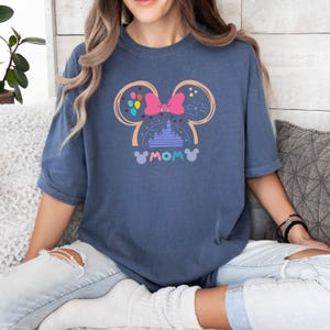 Pode incluir: Uma camiseta azul marinho com um gr&aacute;fico de orelhas do Mickey Mouse com um la&ccedil;o rosa, um castelo e bal&otilde;es. O texto "MOM" est&aacute; dentro das orelhas.