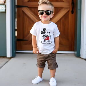 May include: A young boy wearing a white t-shirt with a black and white Mickey Mouse graphic. He is also wearing brown shorts and white sneakers.