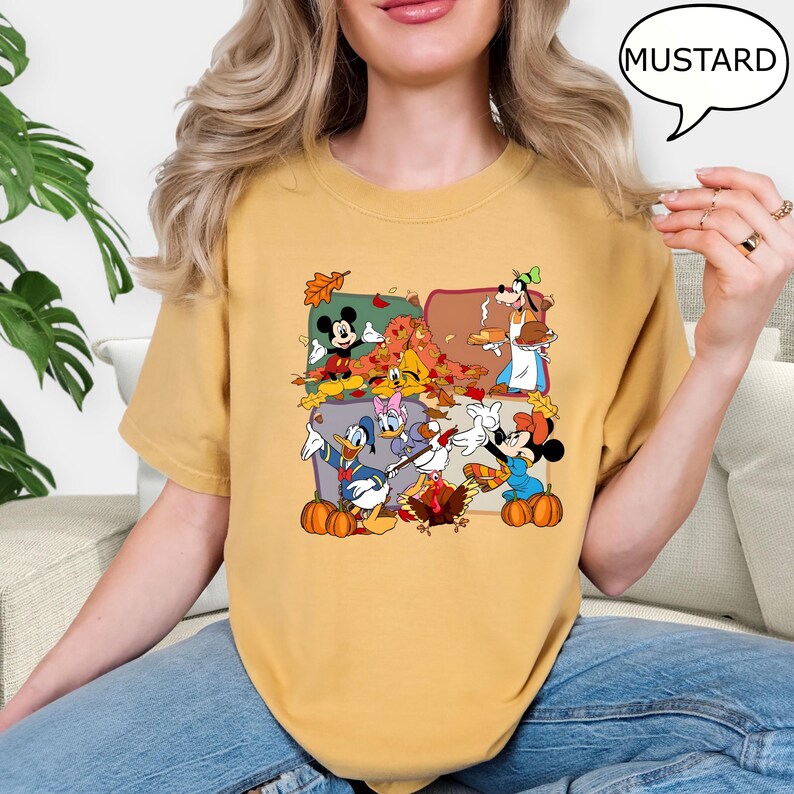 May include: Mustard-colored t-shirt featuring a colorful graphic of cartoon characters celebrating autumn. The graphic includes Mickey and Minnie Mouse, Donald Duck, Goofy, and pumpkins. The word "MUSTARD" is in a speech bubble.