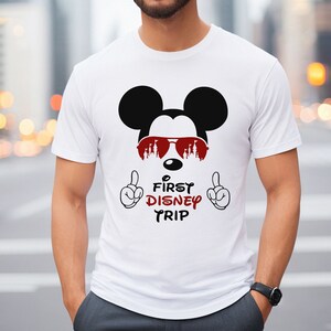 May include: White t-shirt with a black and white image of Mickey Mouse wearing red sunglasses with the text "First Disney Trip" printed on the front.