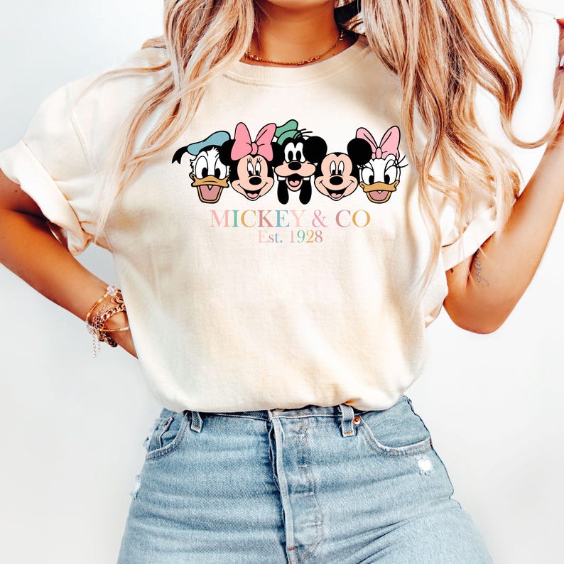 May include: A white t-shirt with a graphic of Mickey Mouse and his friends. The graphic is in a cartoon style and features Mickey Mouse, Minnie Mouse, Donald Duck, Goofy, and Pluto. The text "MICKEY & CO. Est. 1928" is printed below the graphic.