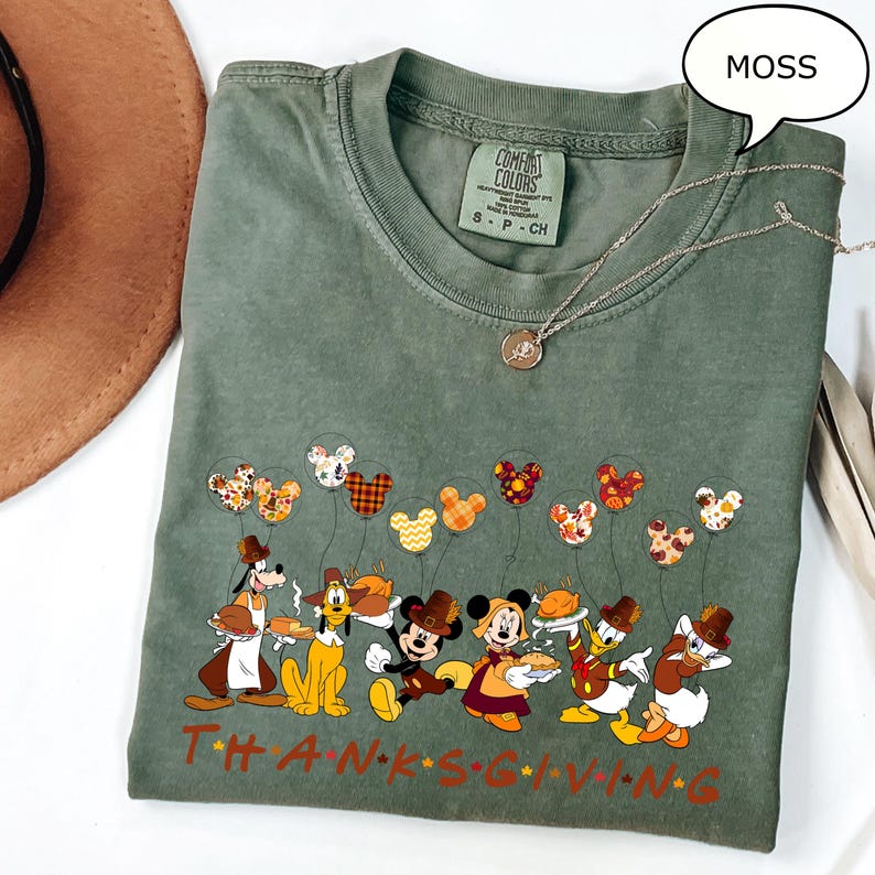 May include: A moss green t-shirt featuring a Thanksgiving-themed graphic with Disney characters. The design includes Mickey Mouse, Donald Duck, and Goofy, holding food and balloons. The word "THANKSGIVING" is printed in orange.