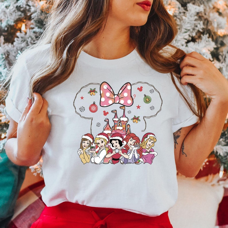 Puede incluir: Camiseta blanca con un dise&ntilde;o navide&ntilde;o con un lazo rosa, un castillo y seis princesas Disney con gorros de Pap&aacute; Noel. Las princesas sostienen objetos navide&ntilde;os.