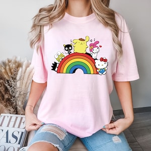 Pode incluir: Uma camiseta rosa com um arco-&iacute;ris e personagens de desenhos animados. Os personagens s&atilde;o um cachorro amarelo, um gato preto, um coelho rosa e um gato branco com um la&ccedil;o vermelho. Os personagens est&atilde;o todos sorrindo e cantando.