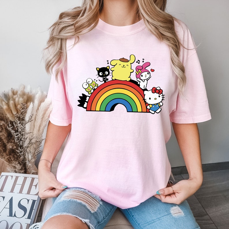 Pode incluir: Uma camiseta rosa com um arco-&iacute;ris e personagens de desenhos animados. Os personagens s&atilde;o um cachorro amarelo, um gato preto, um coelho rosa e um gato branco com um la&ccedil;o vermelho. Os personagens est&atilde;o todos sorrindo e cantando.