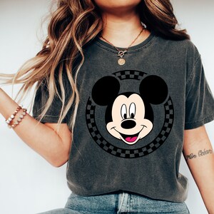 Puede incluir: Una camiseta gris oscura con un c&iacute;rculo a cuadros blanco y negro con la cara de Mickey Mouse. Mickey Mouse est&aacute; sonriendo con la lengua fuera.
