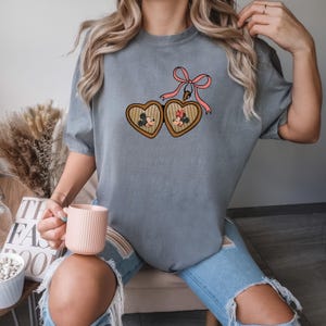 May include: A gray t-shirt with a graphic of Mickey and Minnie Mouse in a heart-shaped locket with a pink bow. The locket is hanging from a chain.