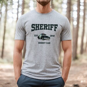 Puede incluir: Una camiseta gris con un gr&aacute;fico negro de un coche de polic&iacute;a de dibujos animados. El texto "SHERIFF EST 1949 DISNEY CAR" est&aacute; impreso en la camiseta.