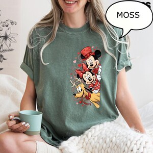 Puede incluir: Camiseta verde musgo con un gr&aacute;fico de Mickey Mouse, Minnie Mouse y Pluto con atuendos de San Valent&iacute;n. Mickey lleva un sombrero de copa rojo, Minnie un lazo rojo y Pluto un collar rojo. La palabra "MOSS" est&aacute; en una burbuja.