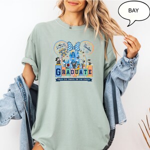 以下が含まれることがあります： ミッキーマウスとその仲間たちが卒業を祝うグラフィックが描かれた薄いブルーのTシャツ。グラフィックには「2025」、「シニア」、「卒業」、「タッセルから城へ」の文字が書かれています。