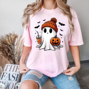 May include: A pink t-shirt featuring a cute cartoon ghost wearing a pumpkin-coloured hat and holding a pumpkin and a cup of orange juice. The ghost has big eyes and a friendly smile. The shirt is ideal for Halloween.