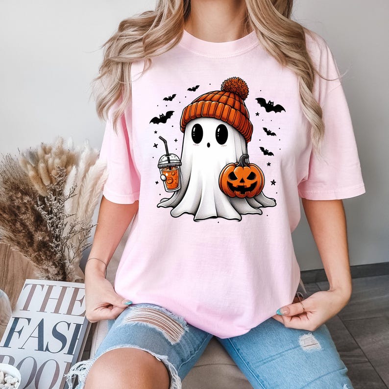 May include: A pink t-shirt featuring a cute cartoon ghost wearing a pumpkin-coloured hat and holding a pumpkin and a cup of orange juice. The ghost has big eyes and a friendly smile. The shirt is ideal for Halloween.