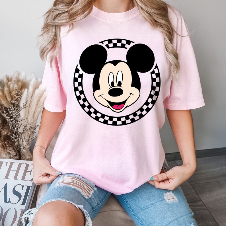 Puede incluir: Una camiseta rosa con un c&iacute;rculo de cuadros negros y blancos alrededor de una imagen de dibujos animados de Mickey Mouse. Mickey Mouse est&aacute; sonriendo con la lengua fuera.