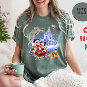 Puede incluir: Una camiseta verde con un dise&ntilde;o navide&ntilde;o que presenta a Mickey Mouse y Minnie Mouse en un trineo con un fondo nevado. El dise&ntilde;o tambi&eacute;n incluye el castillo de Cenicienta y otros personajes de Disney.