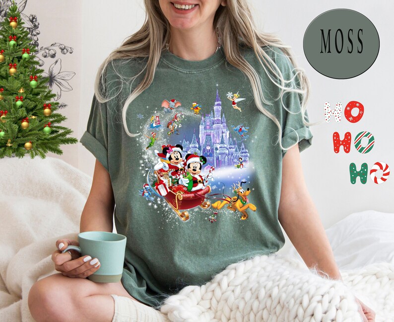 Puede incluir: Una camiseta verde con un dise&ntilde;o navide&ntilde;o que presenta a Mickey Mouse y Minnie Mouse en un trineo con un fondo nevado. El dise&ntilde;o tambi&eacute;n incluye el castillo de Cenicienta y otros personajes de Disney.