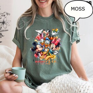 May include: Moss green t-shirt featuring a colorful graphic of Mickey Mouse, Minnie Mouse, Donald Duck, Goofy, and other Disney characters. The graphic includes the text "Mickey & Co. Est 1928".