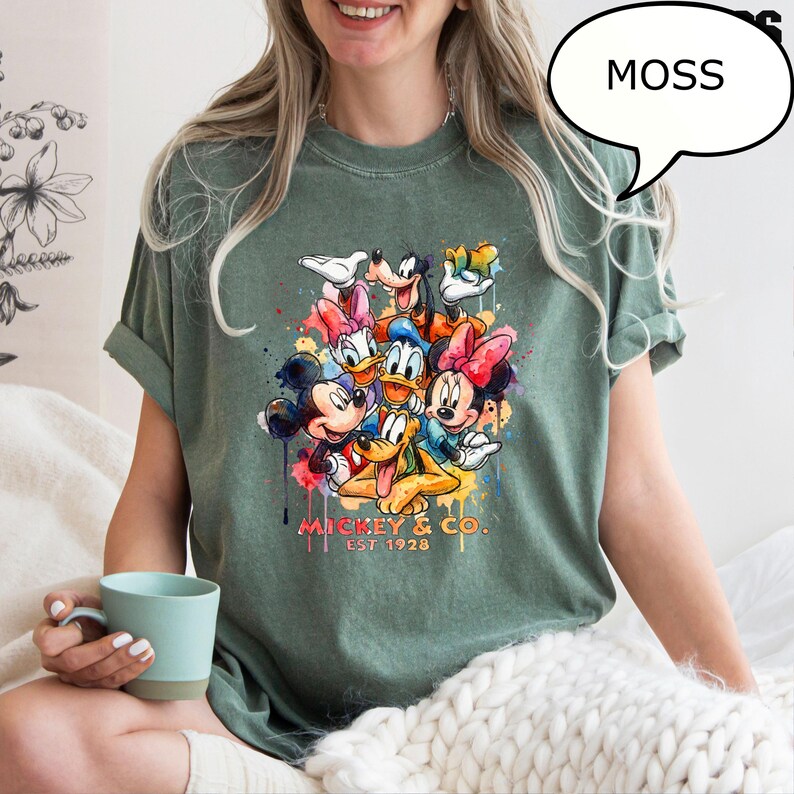 May include: Moss green t-shirt featuring a colorful graphic of Mickey Mouse, Minnie Mouse, Donald Duck, Goofy, and other Disney characters. The graphic includes the text "Mickey & Co. Est 1928".