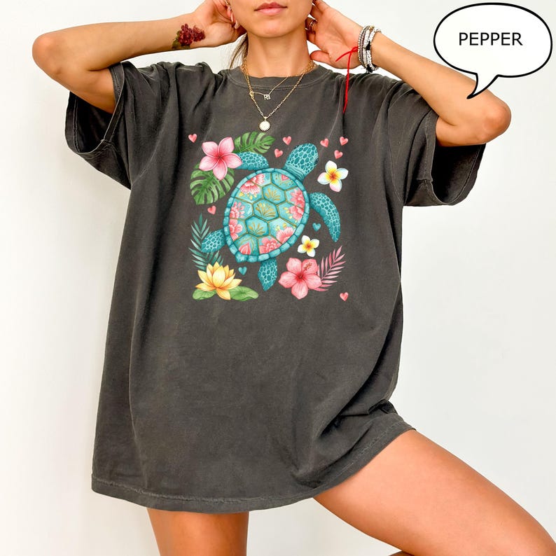 May include: Dark gray t-shirt featuring a colorful sea turtle design. The turtle has a turquoise shell with pink and blue accents, surrounded by pink and yellow flowers, green leaves, and small pink hearts. The word "PEPPER" is in a speech bubble.