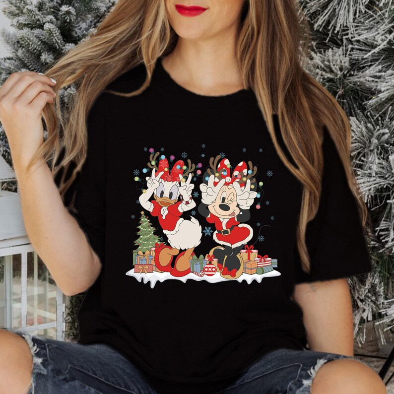 Puede incluir: Camiseta negra con un dise&ntilde;o navide&ntilde;o con Minnie Mouse y Daisy Duck con gorros de Pap&aacute; Noel y sosteniendo regalos. El dise&ntilde;o incluye un peque&ntilde;o &aacute;rbol de Navidad y luces.