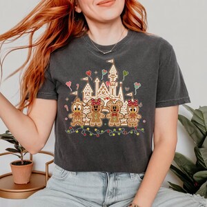 May include: Dark gray t-shirt featuring a gingerbread castle and gingerbread characters. The design includes a castle, gingerbread figures, and colorful string lights. The shirt is a comfortable, casual style.