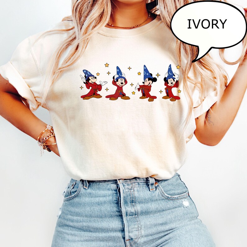 May include: Ivory-colored t-shirt featuring a cartoon design of Mickey Mouse in sorcerer attire. The design includes Mickey in a red robe and blue wizard hat, with gold stars. The word "IVORY" is in a speech bubble.