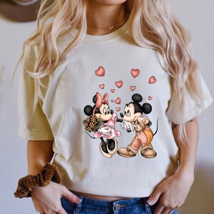 May include: A white t-shirt with a graphic of Mickey Mouse and Minnie Mouse holding hands with hearts floating around them.